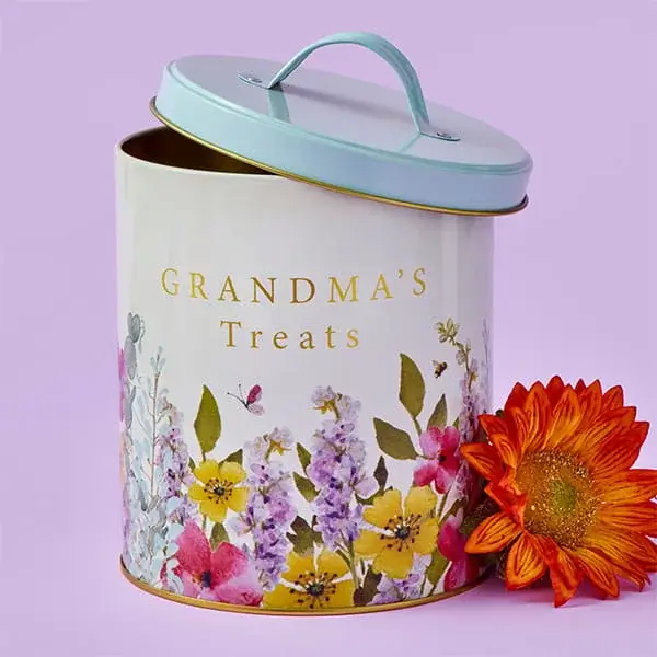 A floral treat tin with a pastel lid, decorated with colourful flowers and the words Grandma’s Treats, shown beside a bright orange flower.